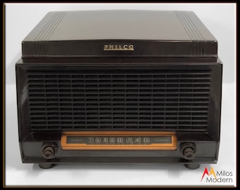 May include: A vintage Philco radio with a black bakelite case and a wood-toned dial. The radio has a grill on the front and two knobs on either side of the dial. The dial has numbers from 55 to 160.