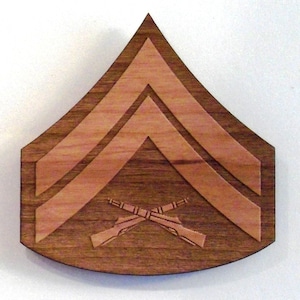 May include: A wooden Sergeant rank insignia with two chevrons and crossed rifles. The insignia is made of light and dark brown wood.
