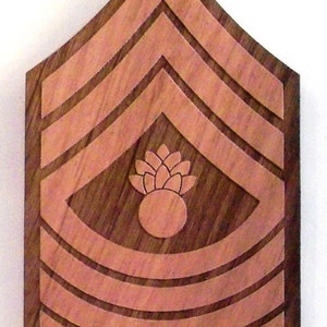 May include: A wooden plaque with a brown background and a light brown design of a military rank insignia. The insignia features three chevrons and a grenade with a pineapple-shaped fuse.