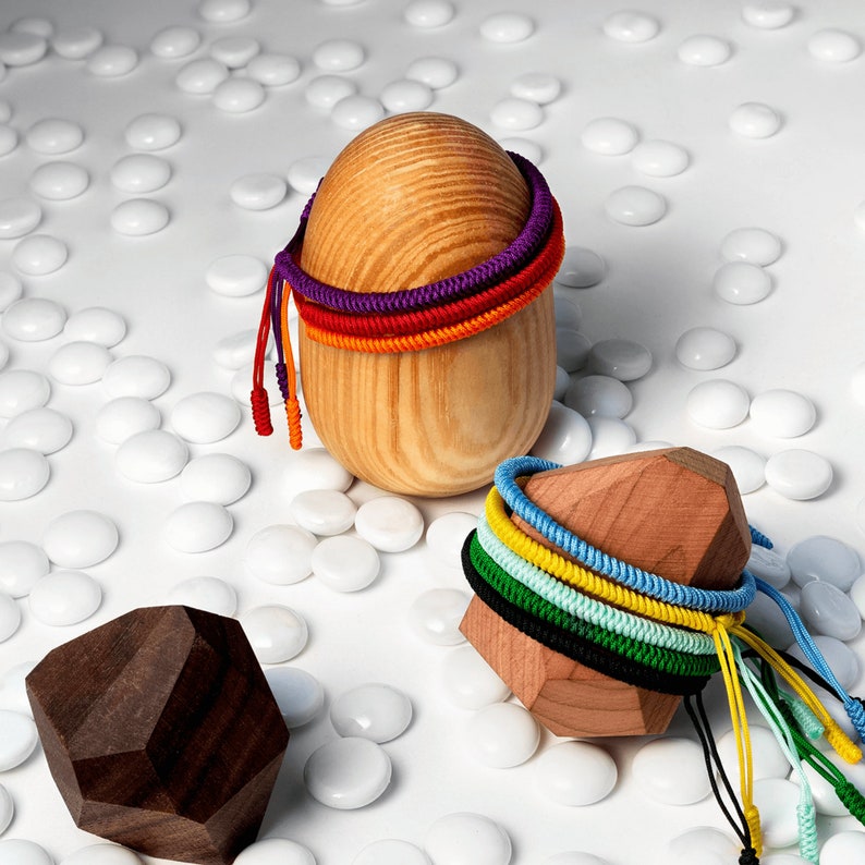 May include: A wooden sphere with colorful string bracelets wrapped around it. The sphere is sitting on a white surface with small white stones scattered around it. There is also a wooden block with colorful string bracelets wrapped around it.