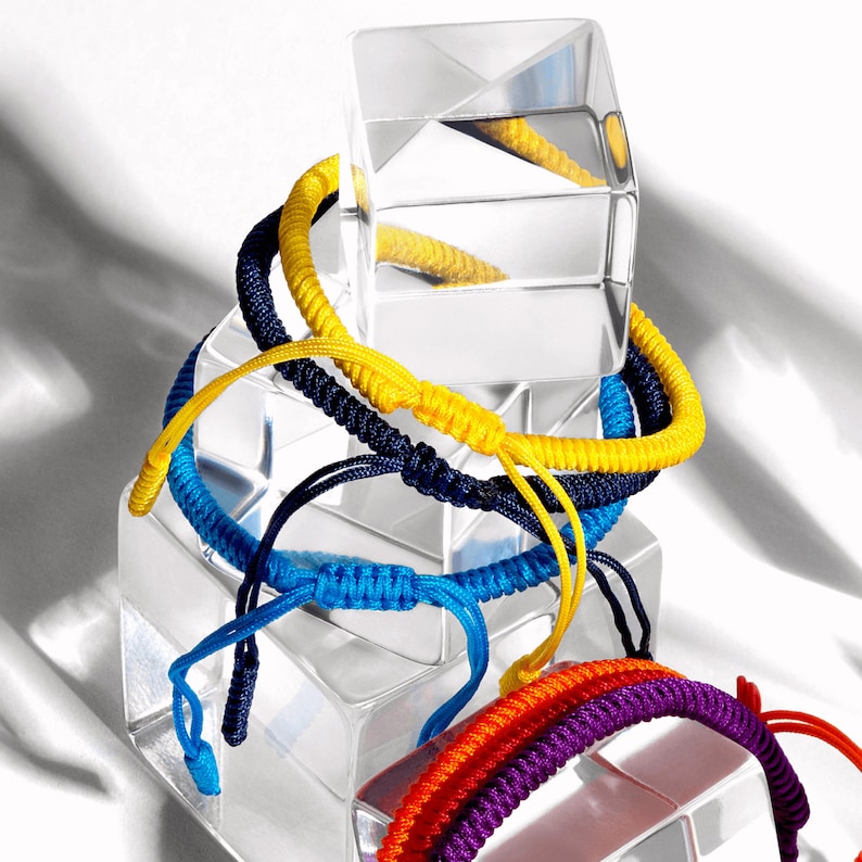 May include: A stack of colorful braided bracelets in yellow, blue, and purple. The bracelets are stacked on top of a clear glass block.