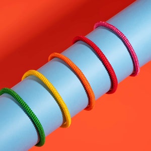 May include: Five colorful braided cords in shades of pink, red, orange, yellow, and green are arranged in a rainbow pattern on a blue surface. The cords are wrapped around the surface, creating a visual effect of a rainbow.