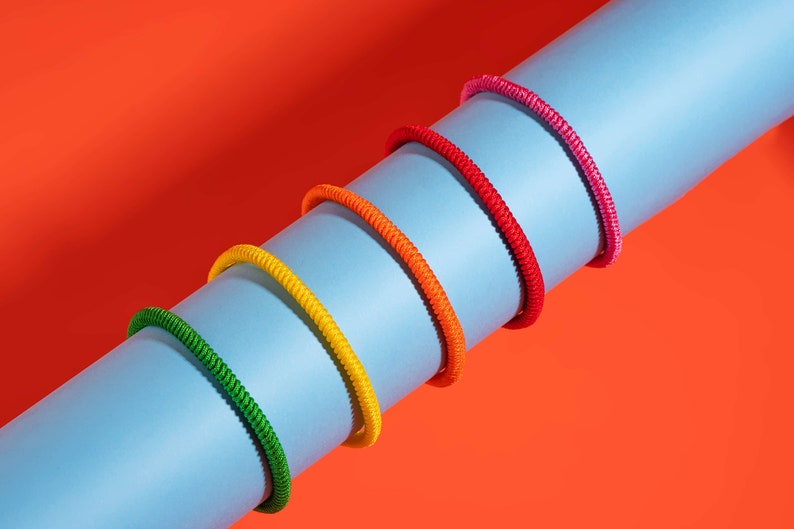 May include: Five colorful braided cords in shades of pink, red, orange, yellow, and green are arranged in a rainbow pattern on a blue surface. The cords are wrapped around the surface, creating a visual effect of a rainbow.