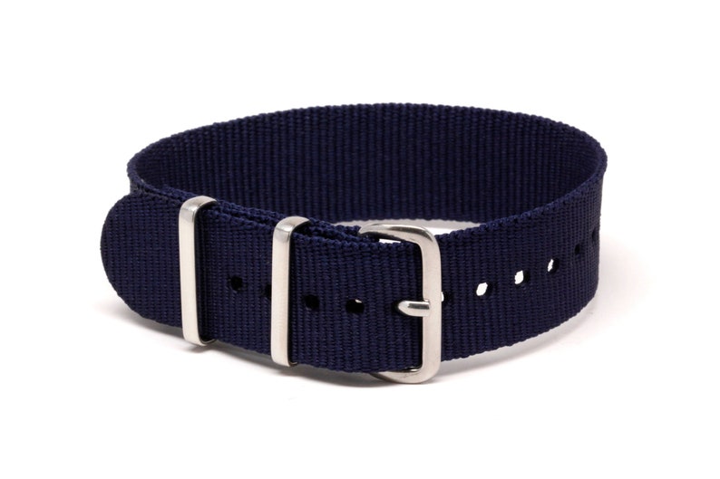 May include: A navy blue watch strap made of woven nylon fabric. The strap features a silver-colored buckle and two silver-colored loops. The strap has several holes for adjustment. The watch strap is designed to be durable and stylish.