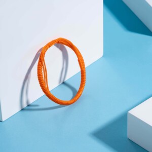 May include: A bright orange braided bracelet on a light blue background. The bracelet is made of a thick cord and is tied in a knot.