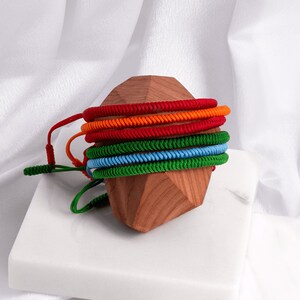 May include: A stack of colorful braided bracelets in red, orange, green, and blue, displayed on a wooden stand.