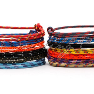 Könnte beinhalten: Ein Stapel bunter Seil-Armbänder, wobei jedes Armband ein anderes Muster aus Rot, Blau, Gelb, Schwarz und Weiß aufweist. Die Armbänder sind in zwei Stapeln angeordnet, einer links und einer rechts.