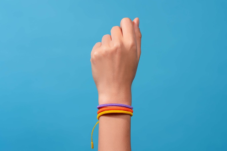 May include: A person's wrist with three colorful bracelets. The bracelets are purple, orange, and yellow.