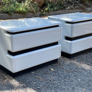 May include: Two white nightstands with a modern design. Each nightstand has two drawers and a black base. The nightstands are made of a glossy material and are placed on a paved surface.