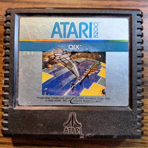 Peut inclure: Une cartouche de jeu vidéo Atari 5200 QIX. La cartouche noire présente une étiquette argentée avec le logo Atari et le titre du jeu. L'étiquette affiche un graphisme coloré d'un vaisseau spatial et de formes géométriques. Informations de copyright visibles.