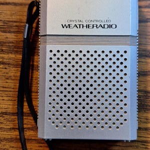 Peut inclure: Radio météo Realistic argenté avec une sangle noire. Le texte "REALISTIC" et "CRYSTAL CONTROLLED WEATHERADIO" est imprimé sur le devant. La zone du haut-parleur présente de nombreux petits trous circulaires.