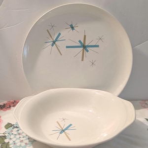 May include: A white ceramic dinner plate and a matching oval serving bowl, both featuring a graphic pattern of light blue and tan starburst shapes with thin black lines. The plate is positioned upright behind the bowl, which has side handles.