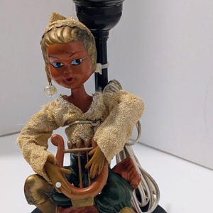 May include: Vintage table lamp featuring a seated figure playing a harp. The lamp has a black base and a black lamp holder. The figure is dressed in gold and green clothing, with a painted face and blue eyes. The lamp is a decorative item.