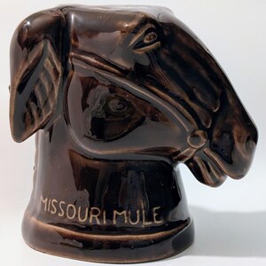 May include: A brown ceramic mug shaped like a mule's head. The mug has detailed features, including the eyes, ears, and bridle. The words "MISSOURI MULE" are inscribed on the base of the mug. The mug is glossy and appears to be vintage.