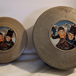May include: Two vintage round decorative trays. The trays have a woven texture and feature a winter scene with two people. The smaller tray is approximately 8 inches in diameter, and the larger tray is about 10 inches in diameter.