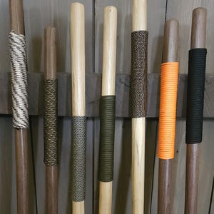 May include: A collection of wooden walking sticks with various grip designs. The grips include woven patterns in white, olive green, and brown, as well as solid orange and black wraps. The sticks are displayed against a wooden background.
