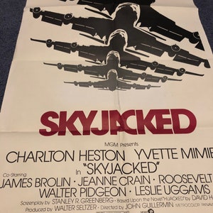 Puede incluir: Un cartel de cine para la película "Skyjacked" protagonizada por Charlton Heston, Yvette Mimieux, James Brolin, Jeanne Crain, Walter Pidgeon y Leslie Uggams. El cartel presenta una silueta de varios aviones volando en formación.