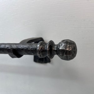 May include: A black metal curtain rod with a hammered finish and a round finial. The rod is mounted to a white wall.