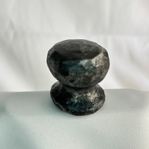 Peut inclure: Un bouton en métal foncé et texturé, avec un dessus arrondi et une base plus petite et évasée. Le bouton a un aspect martelé, avec des imperfections visibles et des variations de couleur gris foncé. Le design général est simple et fonctionnel.