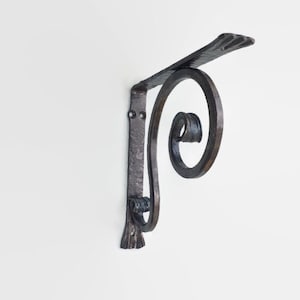 Op de afbeelding: Een zwarte metalen plankbeugel met een decoratief scrollontwerp. De beugel is gemonteerd aan een witte muur.