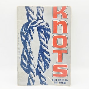 Op de afbeelding: Vintage boekomslag met de titel "Knots" met een grijze achtergrond. De omslag toont een blauw-wit touwknooppatroon en de tekst "Knots" in grote rode letters. De tekst "Boy Scouts of America" staat verticaal aan de linkerkant.
