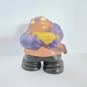 May include: A purple and brown plastic toy figure with a yellow fish in its hand. The figure has a large head with two eyes and a wide mouth.