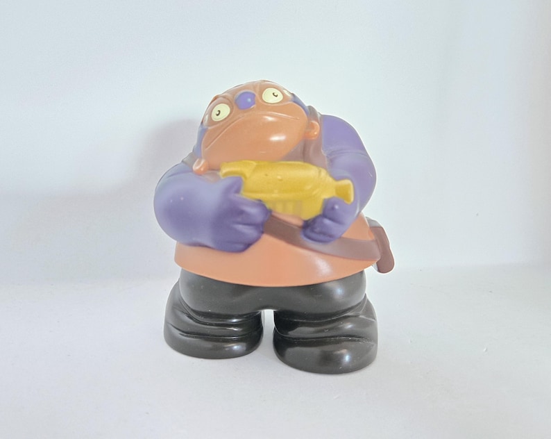 May include: A purple and brown plastic toy figure with a yellow fish in its hand. The figure has a large head with two eyes and a wide mouth.
