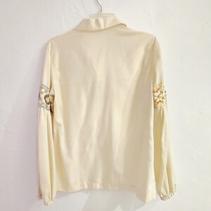 May include: Cream-colored long-sleeve blouse with a collared neckline and decorative floral embroidery on the sleeves. The blouse is hanging on a white wall.