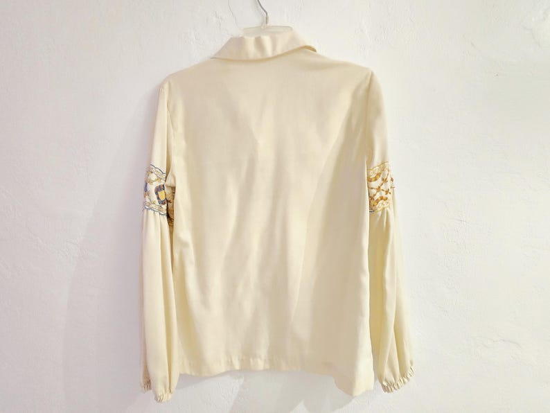 May include: Cream-colored long-sleeve blouse with a collared neckline and decorative floral embroidery on the sleeves. The blouse is hanging on a white wall.