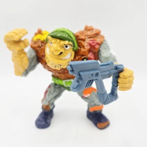Könnte beinhalten: Vintage Actionfigur eines mutierten Charakters mit gelbem Gesicht, orange-braunem Torso und grauen Gliedmaßen. Die Figur hält eine blaue Waffe und trägt einen grünen Helm und Gürtel. Die Figur ist etwa 10 cm groß.