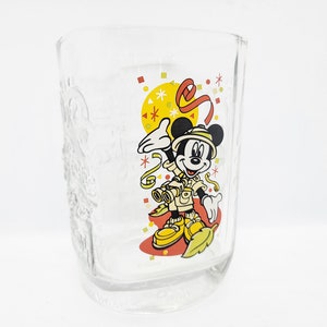 Könnte beinhalten: Ein durchsichtiger Glasbecher mit einem Mickey-Mouse-Motiv. Mickey trägt ein Safari-Outfit und hält ein Fernglas. Der Becher hat ein Konfetti-Design in Gelb, Rot und Weiß.