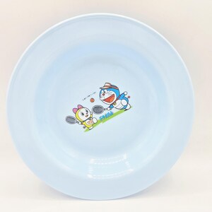 Puede incluir: Un plato redondo azul claro con una ilustraci&oacute;n de dibujos animados de Doraemon y un personaje femenino jugando al tenis. El plato tiene un borde ancho y una superficie lisa, adecuado para servir alimentos.