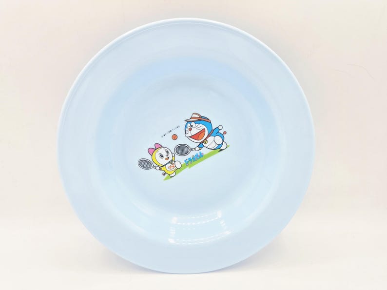 Puede incluir: Un plato redondo azul claro con una ilustraci&oacute;n de dibujos animados de Doraemon y un personaje femenino jugando al tenis. El plato tiene un borde ancho y una superficie lisa, adecuado para servir alimentos.