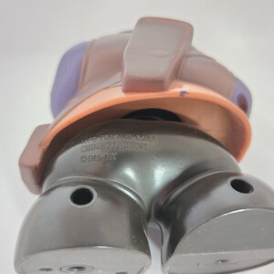 May include: A brown and purple plastic toy figure with a black base. The figure has a large head with a brown hat and a purple face. The base of the figure has the text "WFC TRANSFORMERS CHINA 0711001216 &copy;DISNEY" printed on it.
