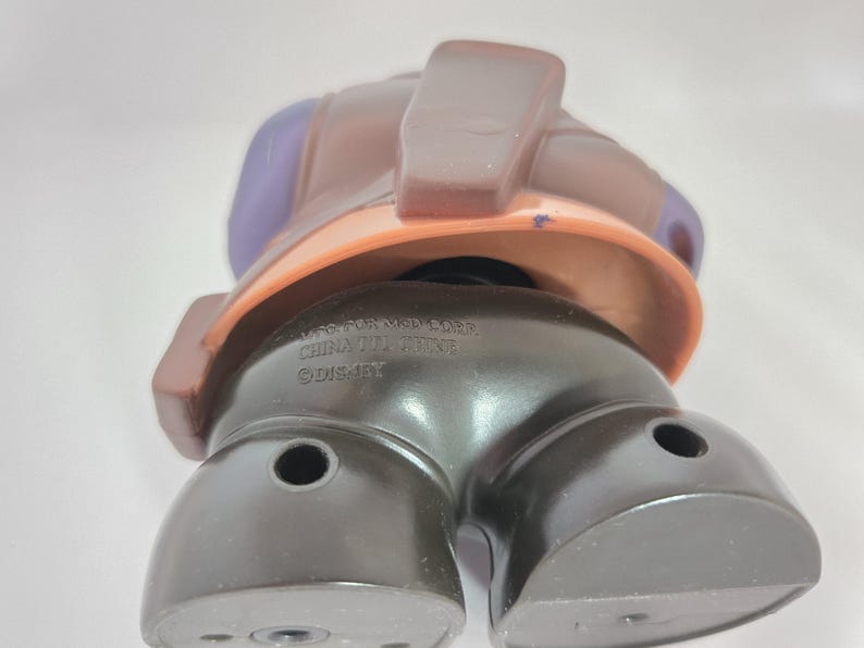 May include: A brown and purple plastic toy figure with a black base. The figure has a large head with a brown hat and a purple face. The base of the figure has the text "WFC TRANSFORMERS CHINA 0711001216 &copy;DISNEY" printed on it.