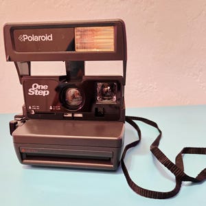 Peut inclure: Appareil photo instantané Polaroid OneStep marron foncé avec une sangle noire. L'appareil photo est équipé d'un flash intégré. Le mot "Polaroid" est imprimé en haut et "One Step" à l'avant.