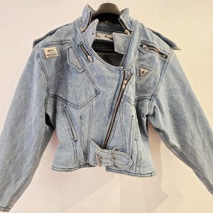 Könnte beinhalten: Hellblaue Jeansjacke mit diagonalem Reißverschluss, mehreren Reißverschlüssen und einem Taillengürtel. Die Jacke hat einen hohen Kragen und dekorative Aufnäher mit silbernen Akzenten. Ein stilvolles, vintage-inspiriertes Oberbekleidungsstück.