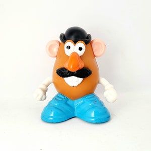 May include: A plastic Mr. Potato Head toy with a brown body, orange nose, black mustache, and a black bowler hat. It has blue shoes, white arms, and pink ears. The toy is a classic children's toy.