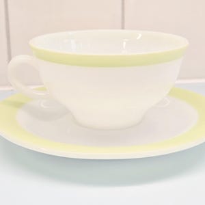 May include: A white teacup and saucer set with a pale green trim. The teacup has a handle and sits on a matching saucer. The set is made of ceramic and is suitable for serving tea or coffee. The set is a classic design.