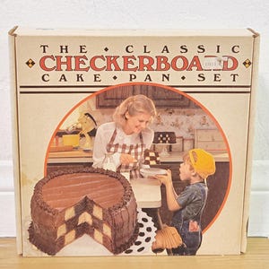 Può includere: Una scatola per un set di teglie per torte a scacchiera. La scatola presenta un'illustrazione di una torta finita con un motivo a scacchiera, e una donna e un bambino in una cucina. Il testo sulla scatola recita "The Classic Checkerboard Cake Pan Set."