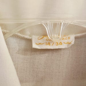 May include: Close-up of a cream-colored garment hanging on a clear plastic hanger. A small tag with gold lettering reads "ON ALS" and "14/34". The fabric has a subtle texture.