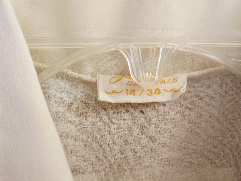May include: Close-up of a cream-colored garment hanging on a clear plastic hanger. A small tag with gold lettering reads "ON ALS" and "14/34". The fabric has a subtle texture.