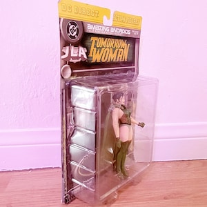 May include: A plastic package containing a green and brown action figure of Tomorrow Woman from the DC Comics Amazing Androids series. The package is yellow and black with the DC Comics logo and the text "JLA" and "Tomorrow Woman".