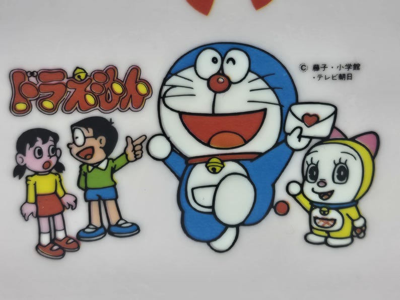 Puede incluir: Ilustraci&oacute;n colorida con personajes de la serie de anime Doraemon. Doraemon, un gato robot azul, es la figura central, rodeado de otros personajes. La imagen incluye texto japon&eacute;s y un dise&ntilde;o en forma de coraz&oacute;n.