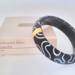 Könnte beinhalten: Ein schwarzes, irisierendes Stoffarmband mit einem goldenen Streifen und einer weißen Schachtel mit der Aufschrift "Avon irisierendes Stoffarmband klein".
