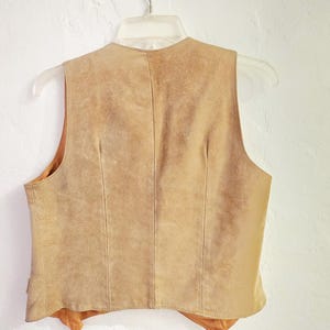 May include: A tan suede vest hangs on a clear plastic hanger against a white wall. The vest has a V-neck and a tailored fit. The back of the vest is visible, showing vertical seams and a soft, textured appearance. The vest is a neutral color.