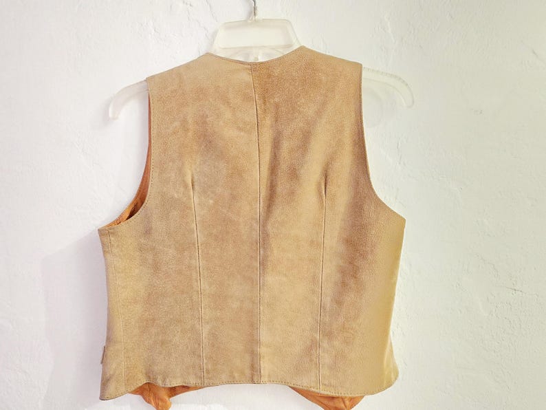 May include: A tan suede vest hangs on a clear plastic hanger against a white wall. The vest has a V-neck and a tailored fit. The back of the vest is visible, showing vertical seams and a soft, textured appearance. The vest is a neutral color.