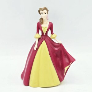 May include: A figurine of a person with brown hair styled in a ponytail, wearing a long dress with a deep red bodice and skirt overlay, and a yellow underskirt. The dress features elbow-length sleeves with yellow cuffs and a yellow neckline.