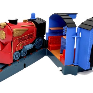 Può includere: Un locomotore giocattolo rosso con un emblema dorato alato e le parole "Hogwarts Express" stampate sul lato. Il treno è su un binario blu e si trova accanto a una stazione ferroviaria giocattolo blu e nera con una piattaforma e un cartello che dice "Platform 9 3/4".