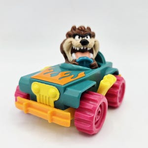 May include: A colorful toy car featuring the Tasmanian Devil character. The car is teal with orange flames, yellow accents, and pink wheels. The Tasmanian Devil figure is brown and is sitting in the driver's seat.